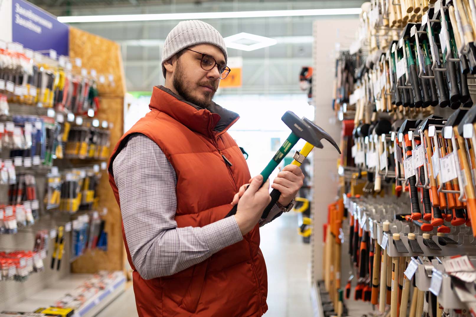 a bearded white man wearing a plaid shirt, orange vest, and gray knit cap compares two hammers in a building materials store