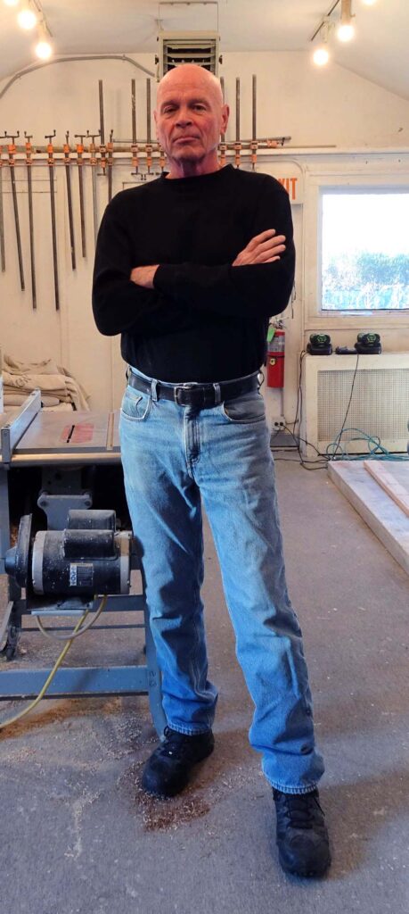 a bald white man wearing a black long-sleeve shirt and blue jeans stands next to a table saw in a garage with a rack of clamps behind him