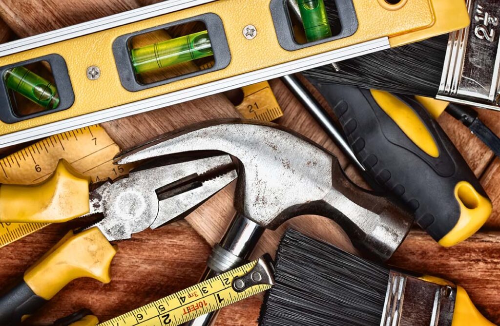 a close up view of manual tools used by contractors, including claw hammer, pliers, bubble level, paintbrushes and ruler on a wooden board background